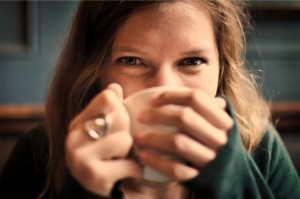 A woman is smiling with her eyes at the camera, since her mouth is drinking from the coffee cup that she's holding up to drink.