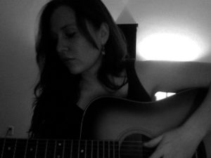 A black & white photo of a woman (me) singing while playing my guitar.