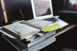 A laptop sits in the background, one side of the home office desk. On the other side, in the foreground, a stack of notebooks and reading books are piled high, with a handwritten journal open-faced on top of the pile.