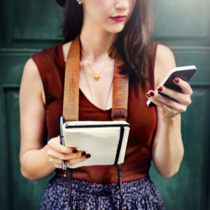 A young woman stands with a notebook in one hand and her cell phone in the other. She's looking at something on her cell phone.