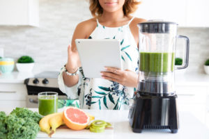 Woman in kitchen smiles at the tablet in her hand. A blender filled with green juice and more veggies, sits on the counter.