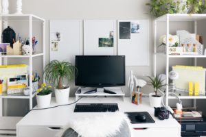 A desktop computer sits at the center of a vacated desk. Office supplies and shelves are all around but no people...