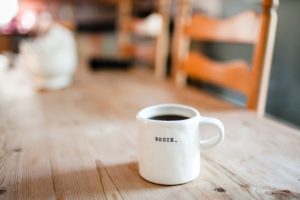 A cup of coffee with the word "begin" painted on it, sits in the middle of an empty wooden table.