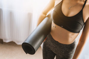 A woman stands alone with a new yoga mat underneath her arm. She's dressed and ready for class.