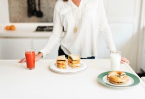 A woman stands in her kitchen with a breakfast sandwich in front of her and a veggie juice on the side. Another breakfast sandwich is made for someone joining her, along with a glass of milk.