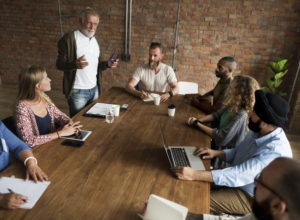 One man is standing in front of a room of about a dozen co-workers at a conference table. He's speaking to them directly.