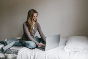 A young women is sitting on her bed, typing on her laptop.