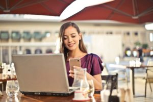 A lovely young, smiling woman is sitting at her laptop, while simultaneously on her cell phone, at an outdoor cafe