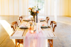 A long wooden table with wood & gold chairs, a blush pink table runner lay down the middle and a beautiful floral centerpiece on top of the table