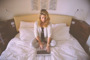A woman sits on her bed alone, typing on her laptop.