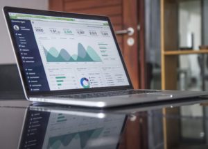 An open laptop sits on a glass tabletop. The webpage that's showing is one with graphs and charts, like an data analytics report for marketing metrics.