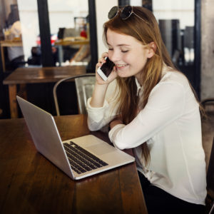 A young woman is on her cell phone, smiling, while looking at her laptop, while sitting in a coffee shop.