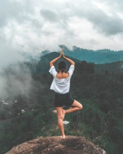A woman does a yoga pose at the top of a mountain, over looking a cloudy mountain range