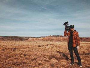 A film cameraman stands in the desert, holding his camera on his right shoulder.