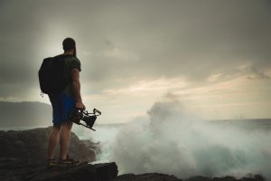 A man stand upon a cliff overlooking the ocean and a huge storm off in the distance. It seems as though he's packed up to leave just in time, before the storm hits the shore (and him).