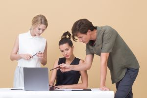 A man and a woman stand behind another woman who is sitting at a laptop. The man is pointing something out to the women about what's on the screen