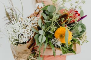 A woman in the background is holding a massive bouquet of wildflowers wrapped in brown paper, holding them up to the camera